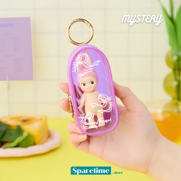 A hand holds a small oval keychain or bag-shaped accessory with a gold ring, featuring a tiny doll inside a translucent lavender case with white line-art graphics. The background is a softly blurred pastel kitchen scene with the words &