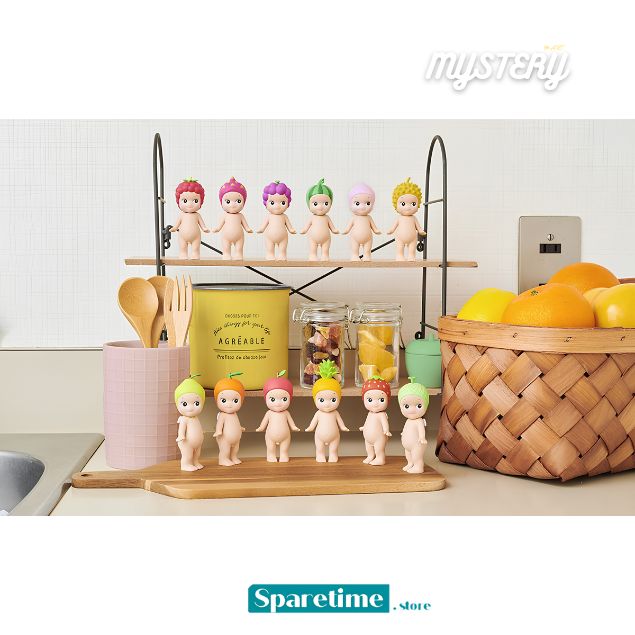 A set of six small nude baby dolls with colorful hair stands on a black metal two-tier shelf and a wooden tray in a kitchen setting, alongside a basket of oranges, a pink cup with utensils, and jars.